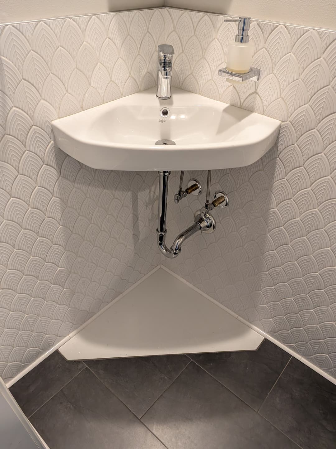 Close-up of corner sink installation in a remodeled half bathroom with white textured tile and chrome plumbing
